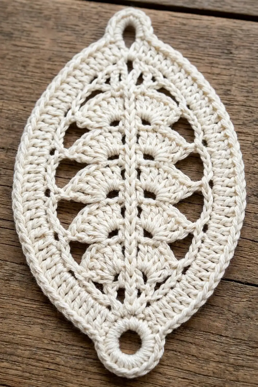 Cream-colored crocheted leaf-shaped ornament with symmetrical lace openwork design and hanging loop on wood backdrop.