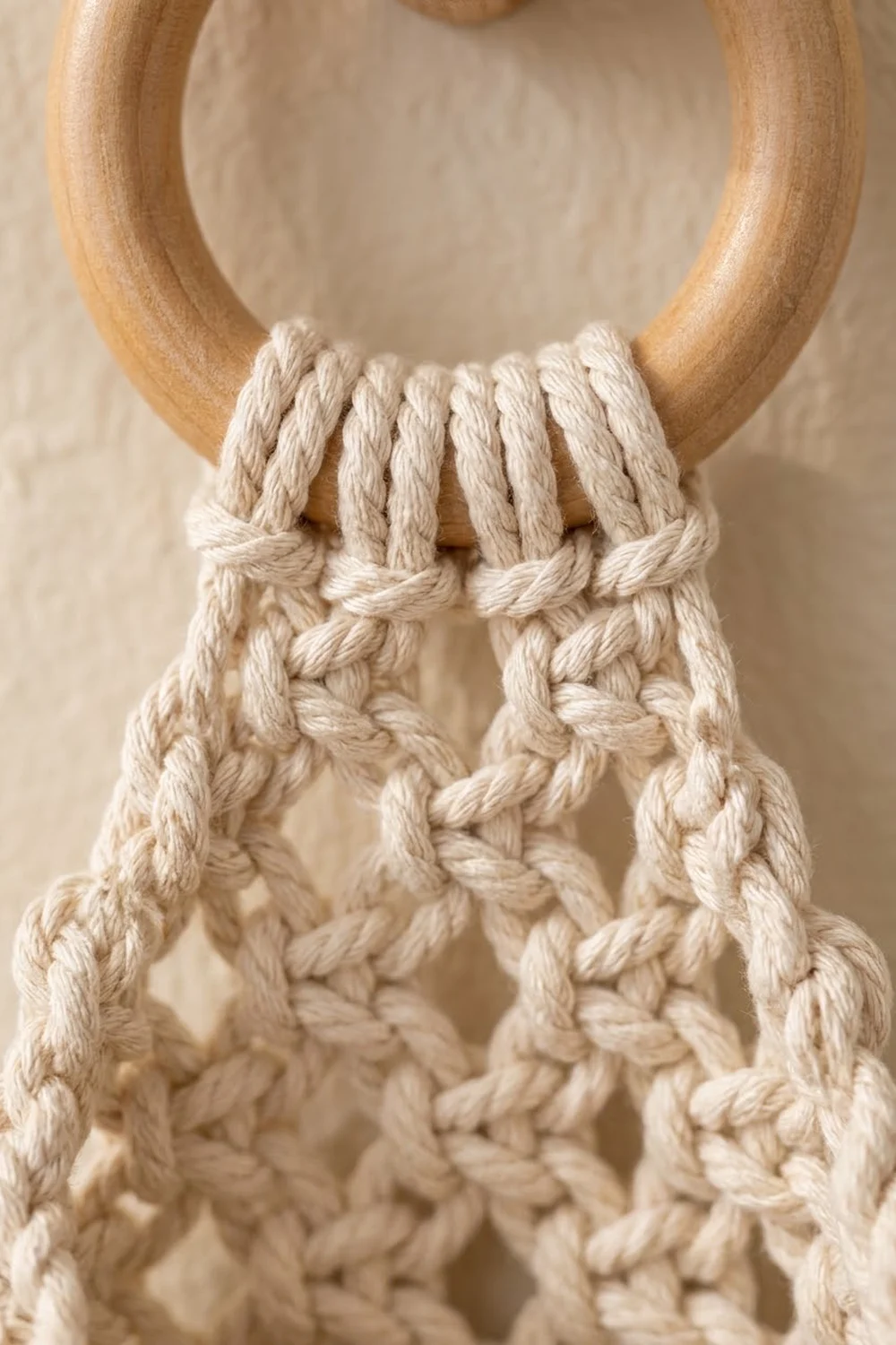 Cream macramé rope with twisted knots and woven patterns, suspended from a natural wooden ring against a soft beige background.