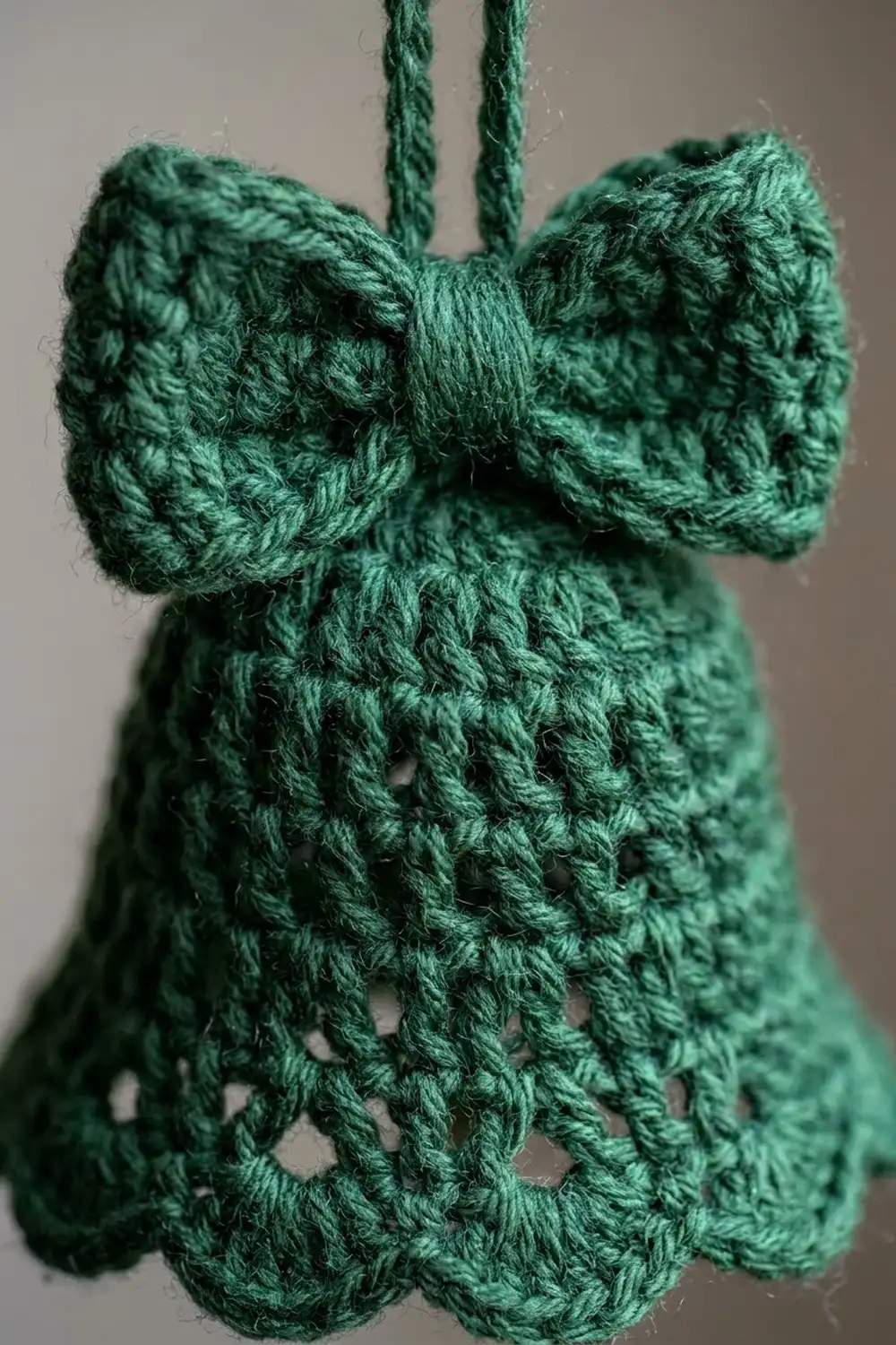 Close-up of a green crocheted ornament with a bow and hanging strings against a blurred gray background.