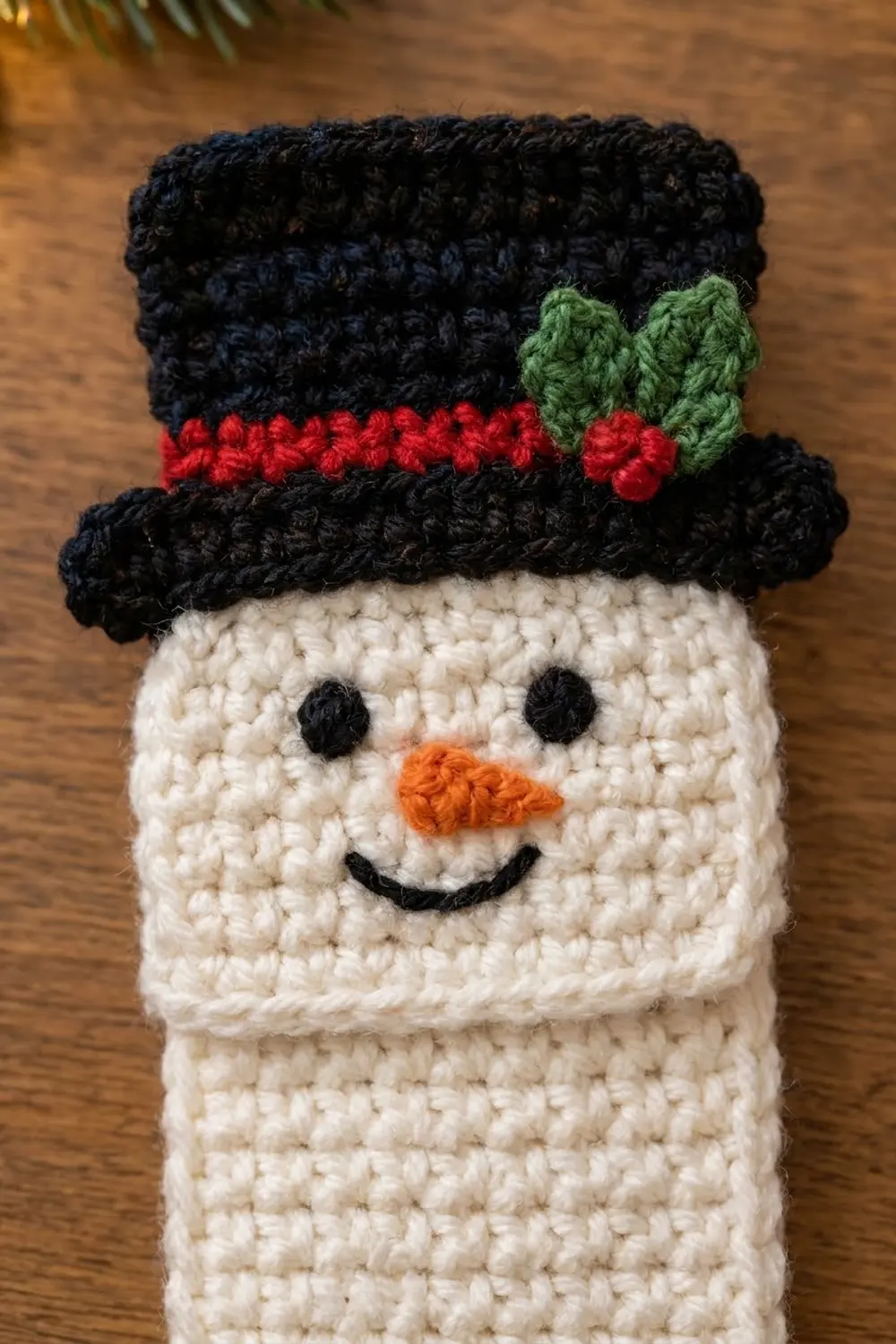 Close-up of crocheted snowman with cream-colored head, black top hat trimmed with red and green holly, orange nose, on wood.