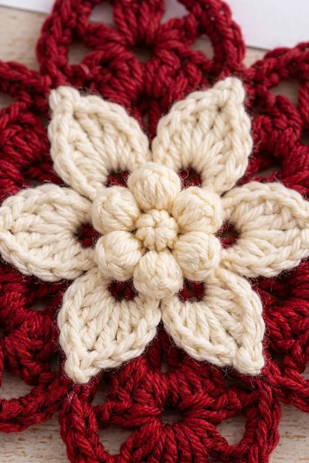 Cream-colored crocheted flower with six textured petals centered on a deep red yarn background.