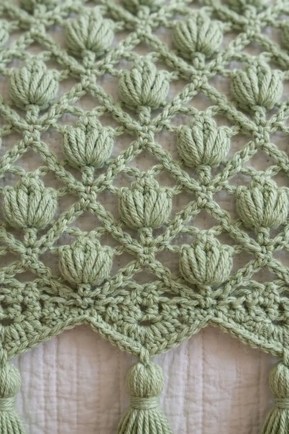 Sage green crochet with pom-pom bobbles in a latticed pattern, featuring a scalloped edge draped over white linen.