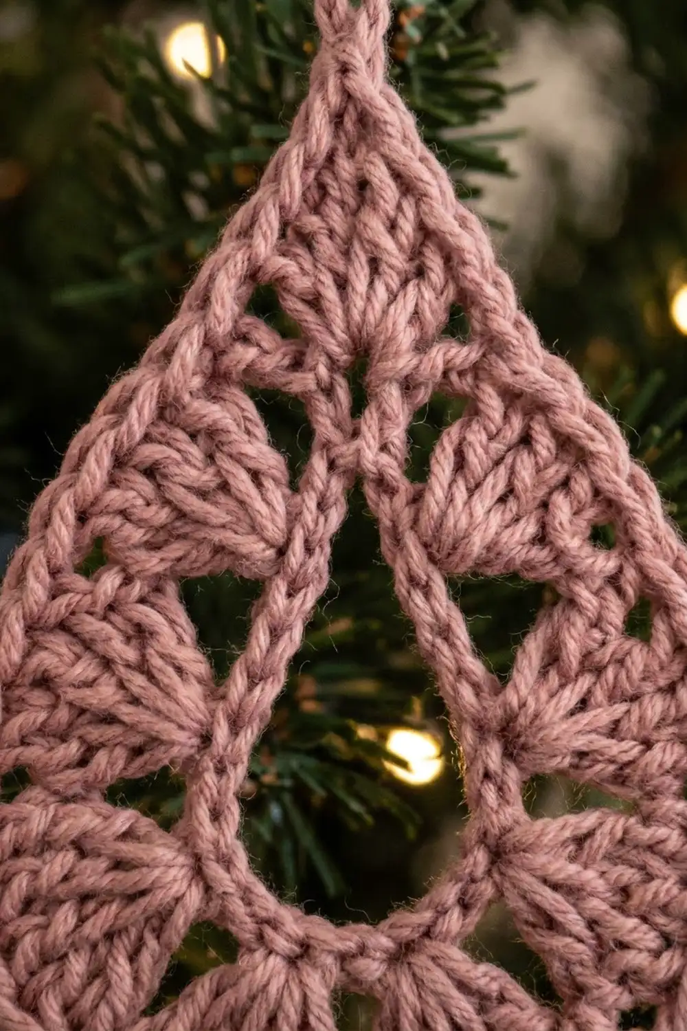 A mauve crocheted teardrop ornament hangs among evergreen branches and warm-glowing lights.