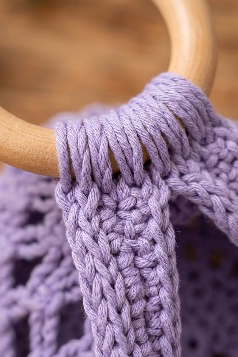 Close-up of light purple crocheted fabric held in hands, displaying intricate stitch detail against a soft blurred background.