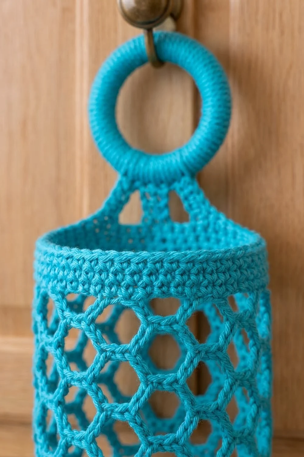 Turquoise crocheted drawstring bag with open lattice pattern and looped handle hangs from a metal hook on wood background.