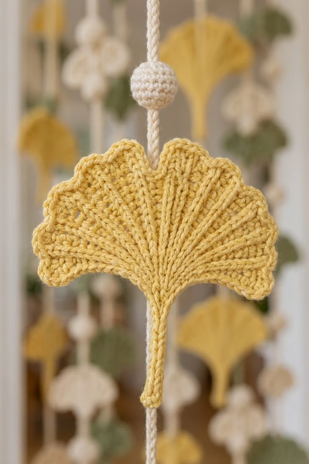 Golden crocheted leaf hanging from cream rope with a small white crochet ball, surrounded by soft-focused ornaments.
