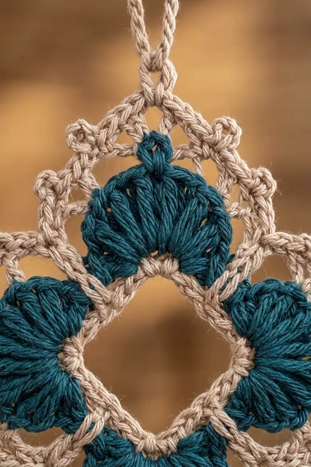 Macramé ornament with natural twine rope and teal yarn tassels hanging against a blurred tan background.