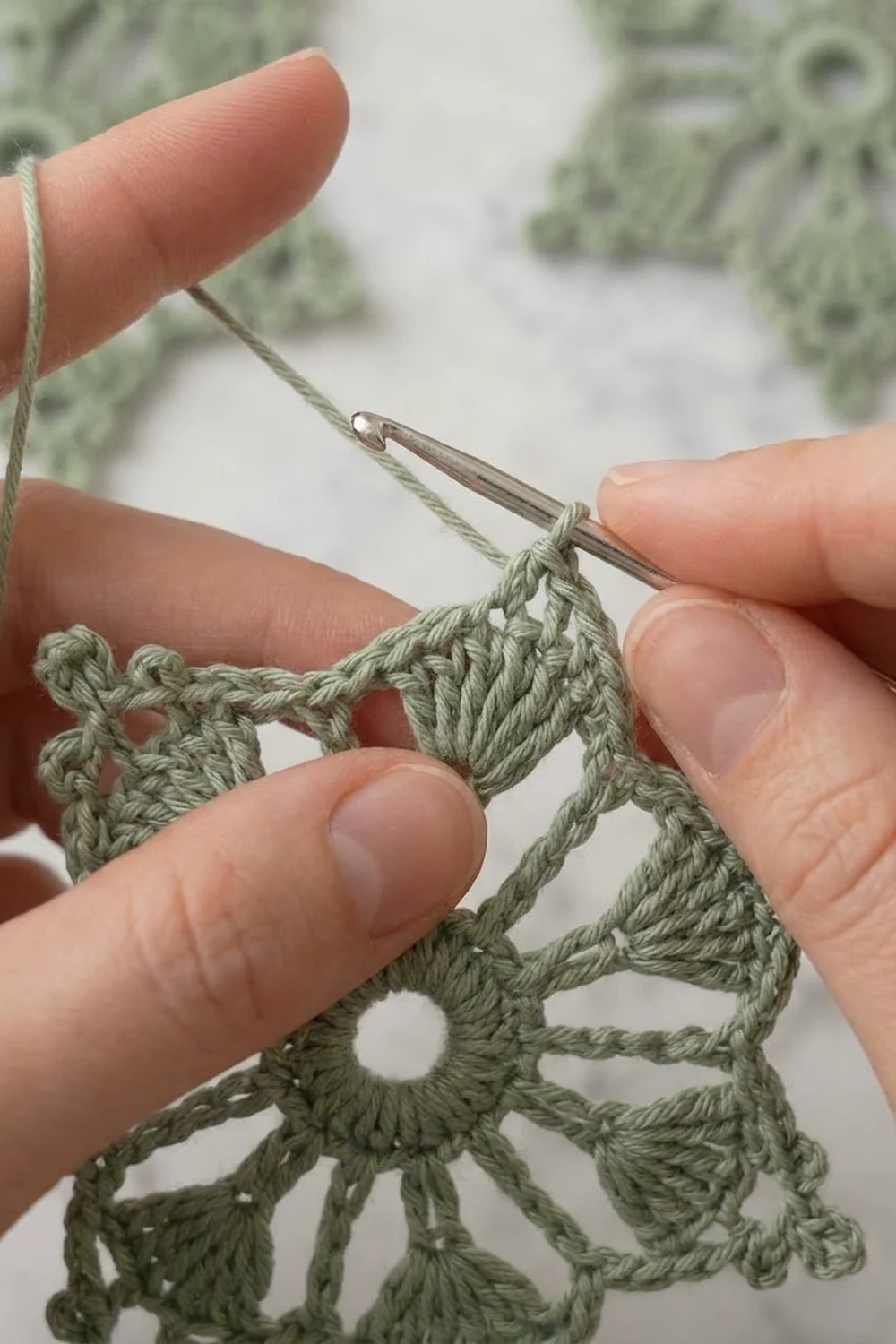 Close-up of hands crocheting sage green lace with a metal crochet hook against a white background.