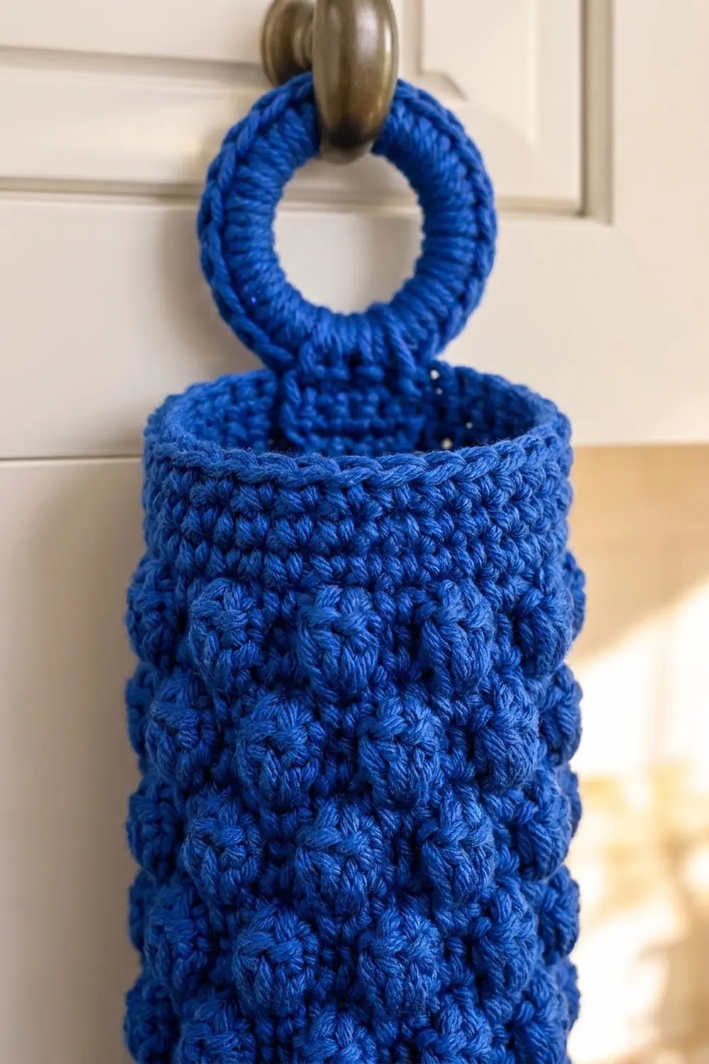 Blue crocheted pouch with textured bobble stitch hanging from a metal rod against cream wall.