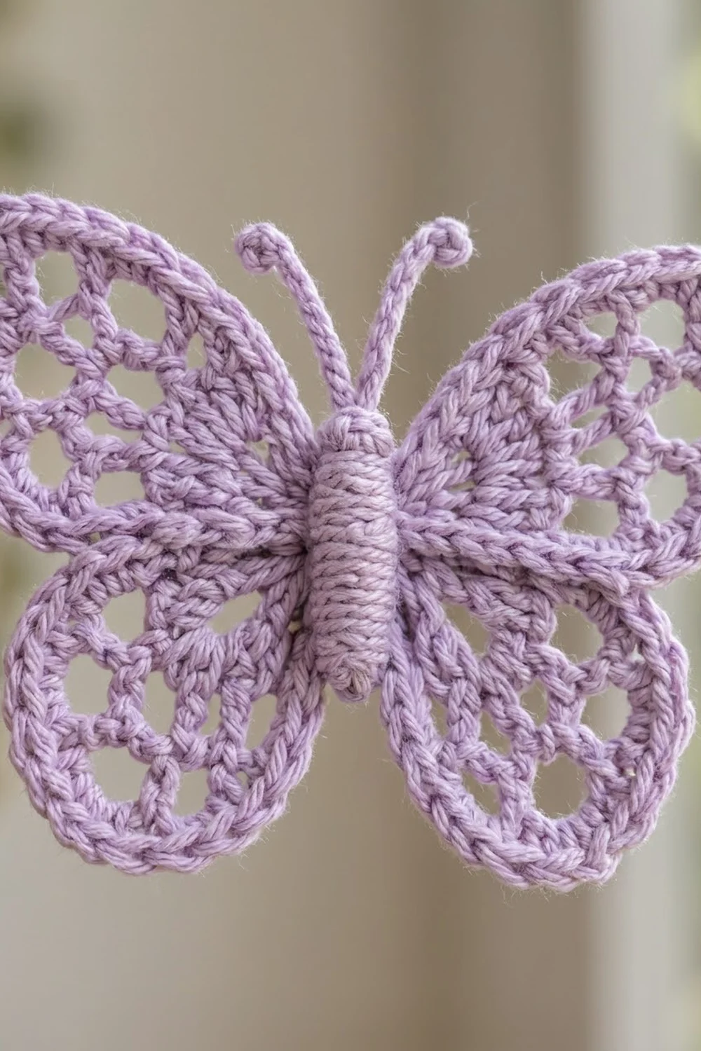 Delicate lavender crochet butterfly with lacy openwork wings and curled antennae on a neutral blurred background.