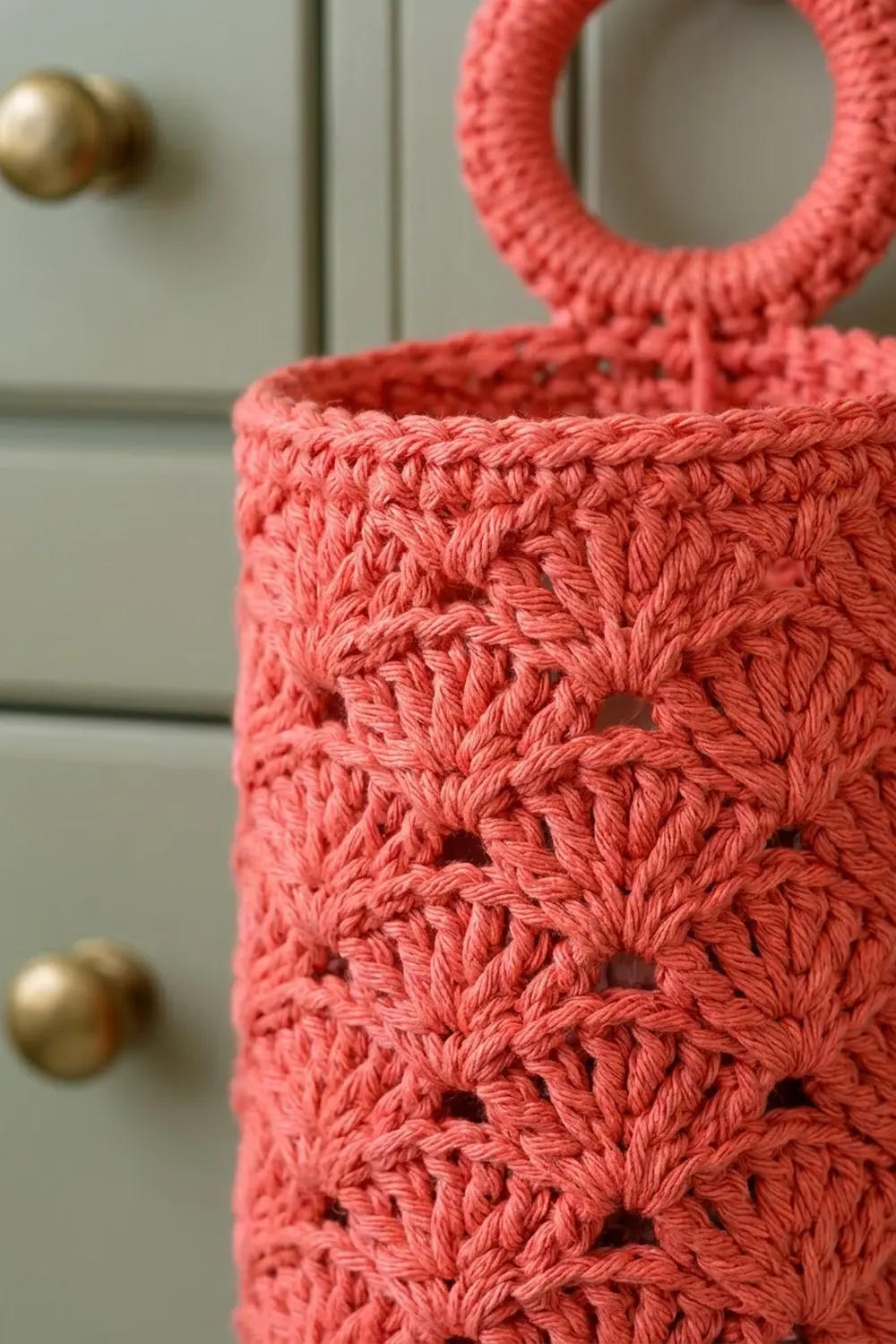 Close-up of coral-pink crocheted tote with open lace pattern and handles against light gray wall with brass hardware.