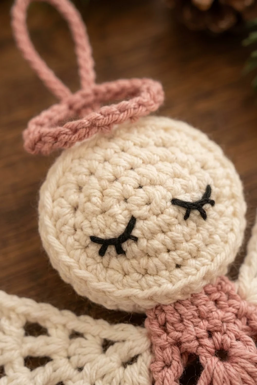 A cream-colored crocheted bunny ornament with closed eyes and pink ears against a blurred brown background.