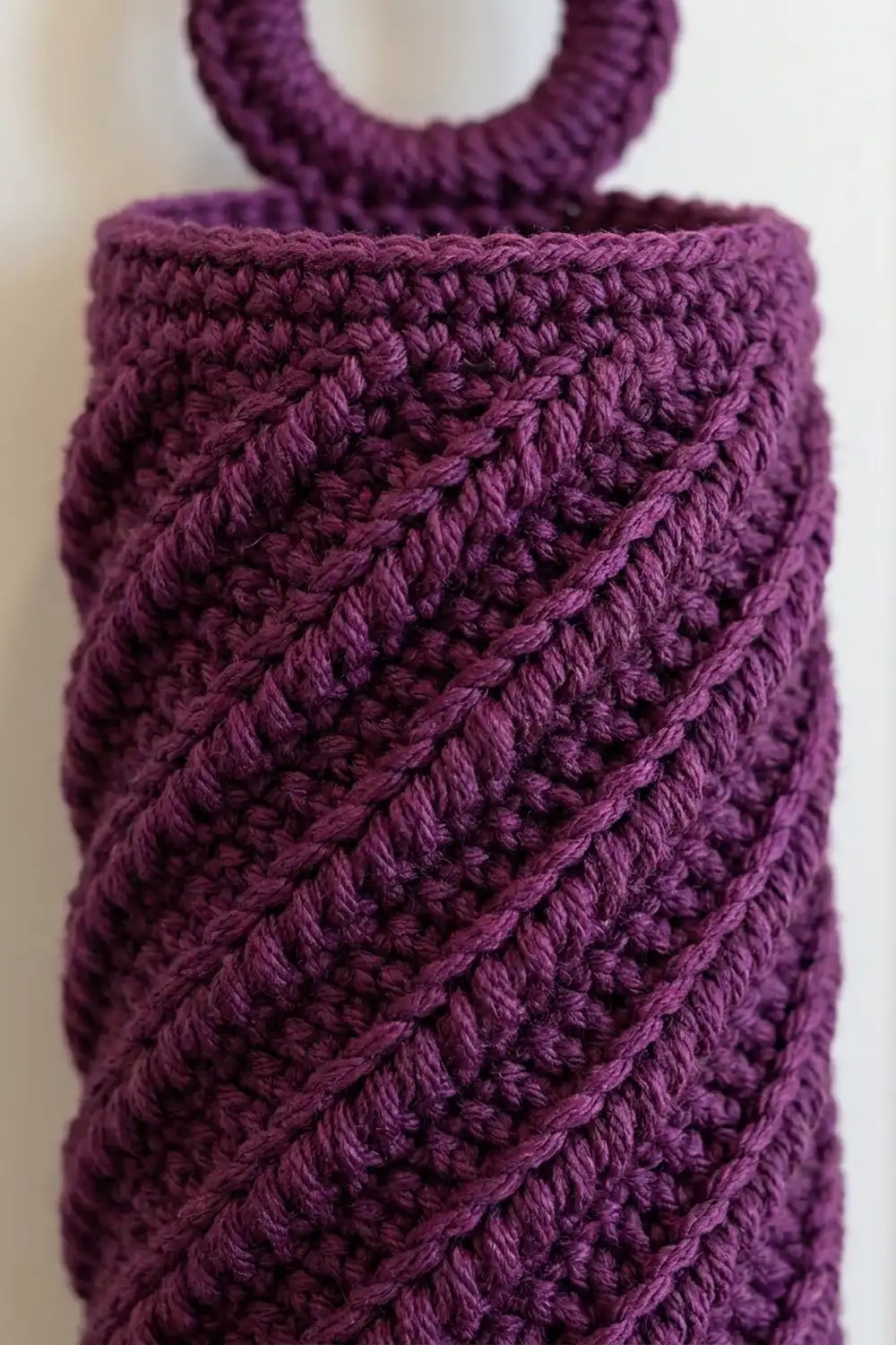 Deep purple crocheted bag with diagonal textured stitches and handles, photographed against a warm, neutral background.