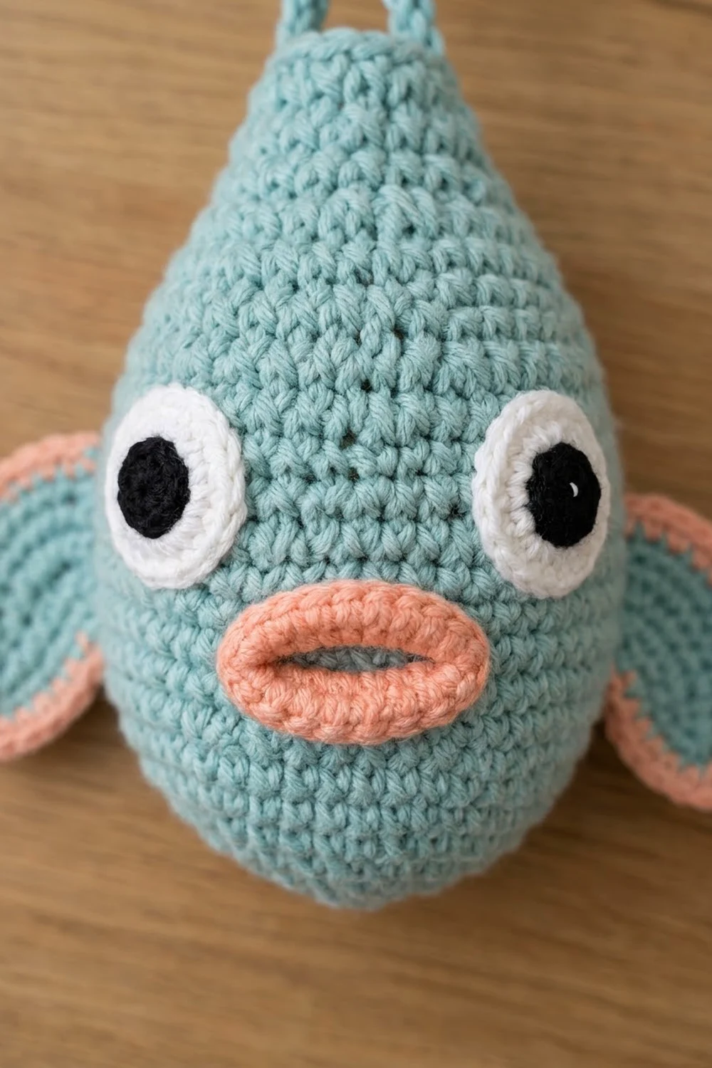 Handmade light-blue crochet bird with textured stitches, white-rimmed eyes, and coral mouth on wood.