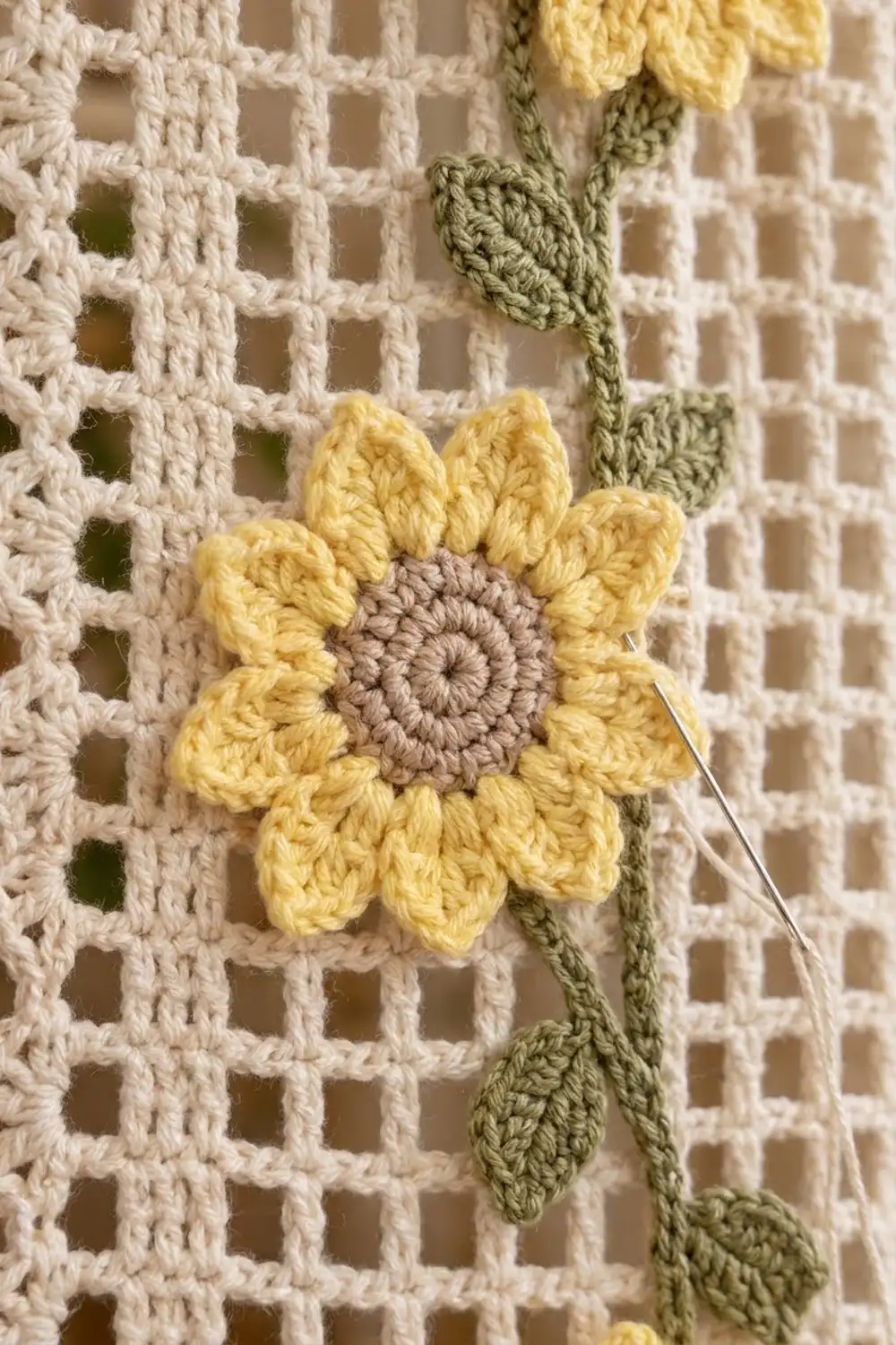 Yellow crocheted flower with taupe center on cream openwork background with green stems and leaves.