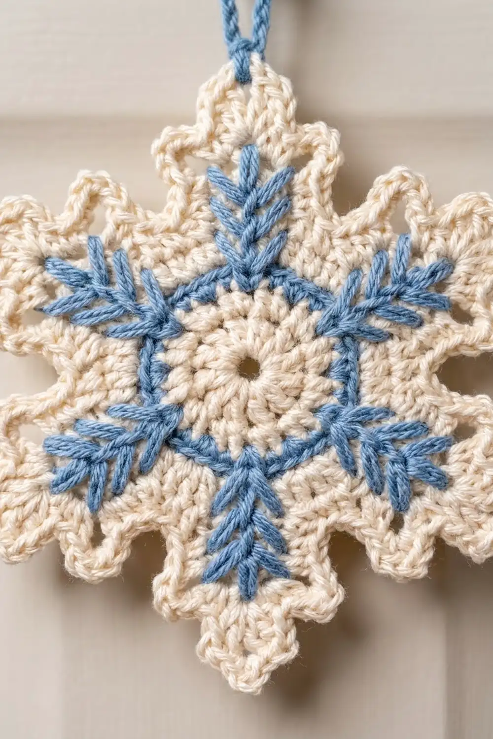 Crocheted cream snowflake with blue embroidered star detail, hanging from a blue yarn loop on a neutral background.
