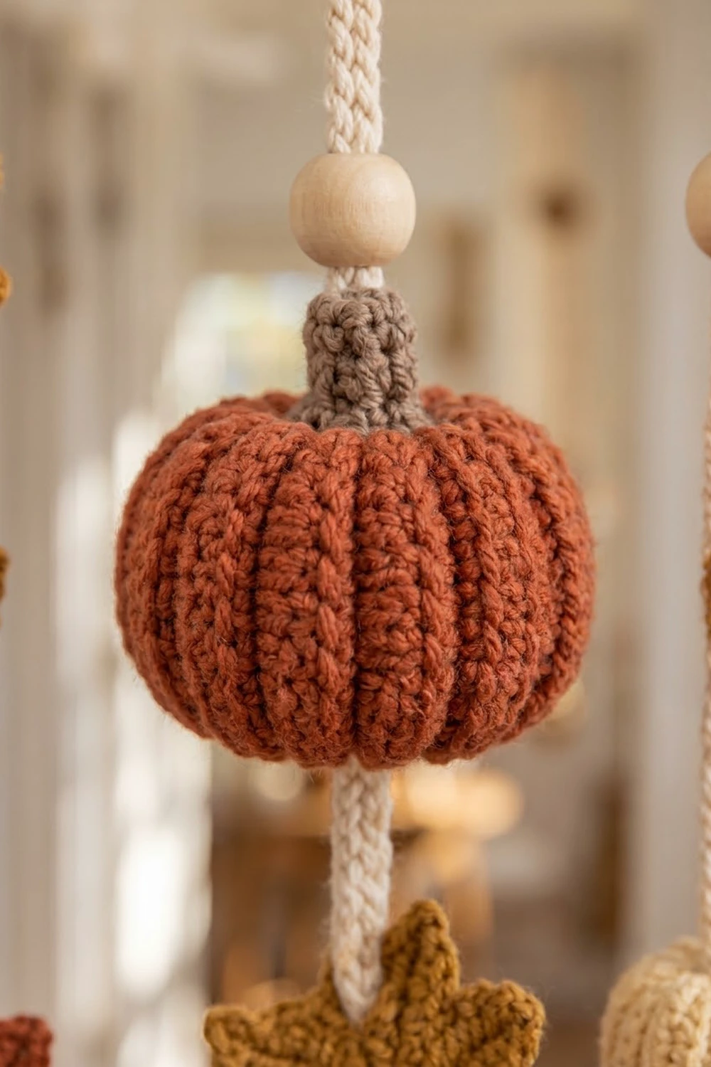 Crocheted rust-colored pumpkin ornament with vertical ribbed texture, suspended by cream rope and natural wood bead, against blurred warm background.