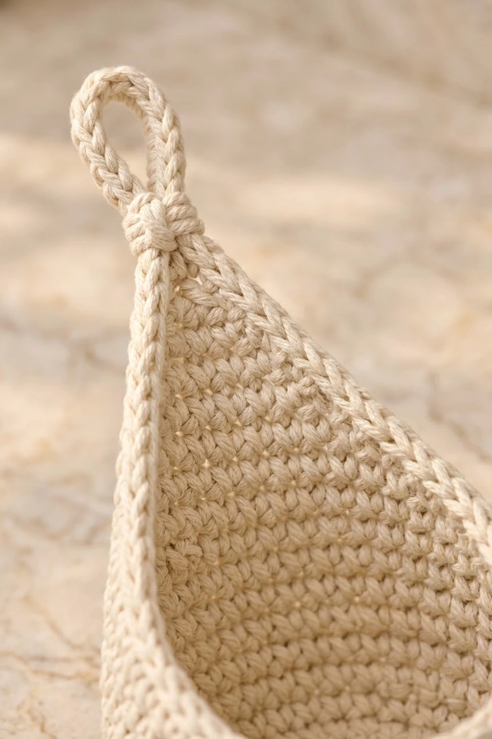 Cream-colored crocheted tote with raised popcorn stitch and braided handle detail on neutral sand-toned background.