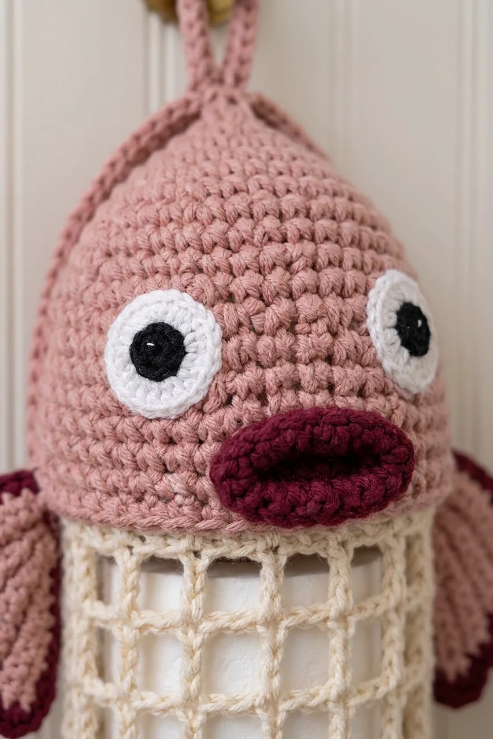 Crocheted pink ice cream cone character with white eyes, burgundy mouth, and cream waffle cone body against a blurred background.
