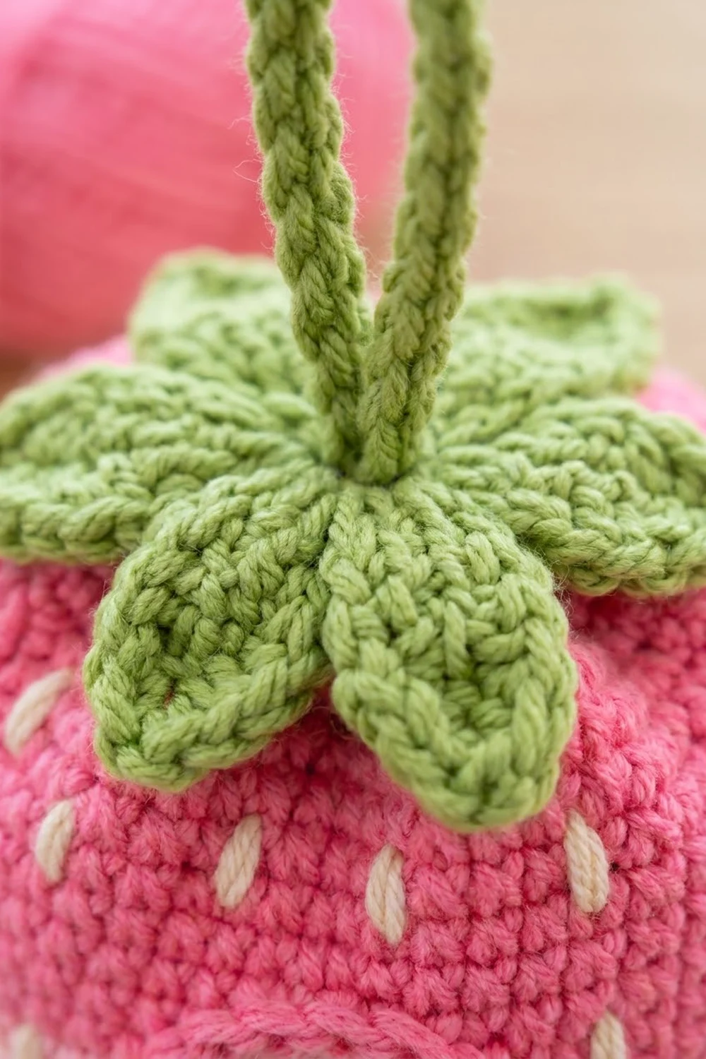 Close-up of a pink crocheted strawberry with sage green leaves and white embroidered seed details.