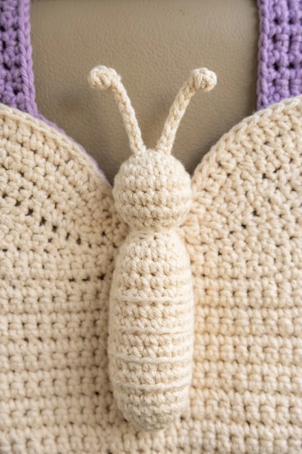 Crocheted cream butterfly with segmented abdomen, long antennae, and visible handmade stitches, photographed in close-up against purple-trimmed fabric.