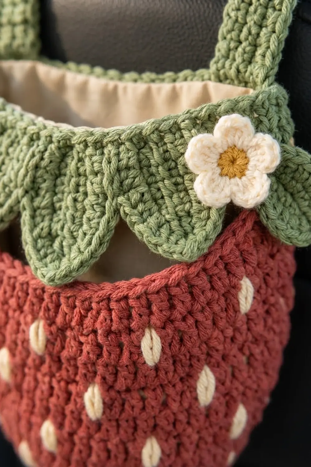 Close-up of crocheted fabric detail showing sage-green texture, white daisy with gold center, and rust-red base with cream buttons.