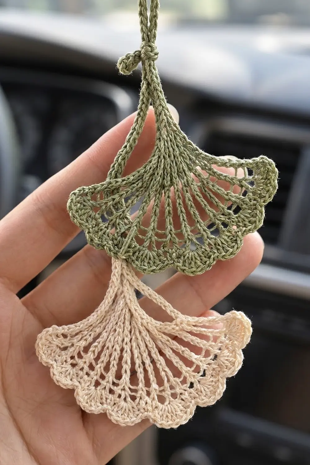 Two crocheted ginkgo-leaf ornaments in sage green and cream with delicate openwork, held in palm.