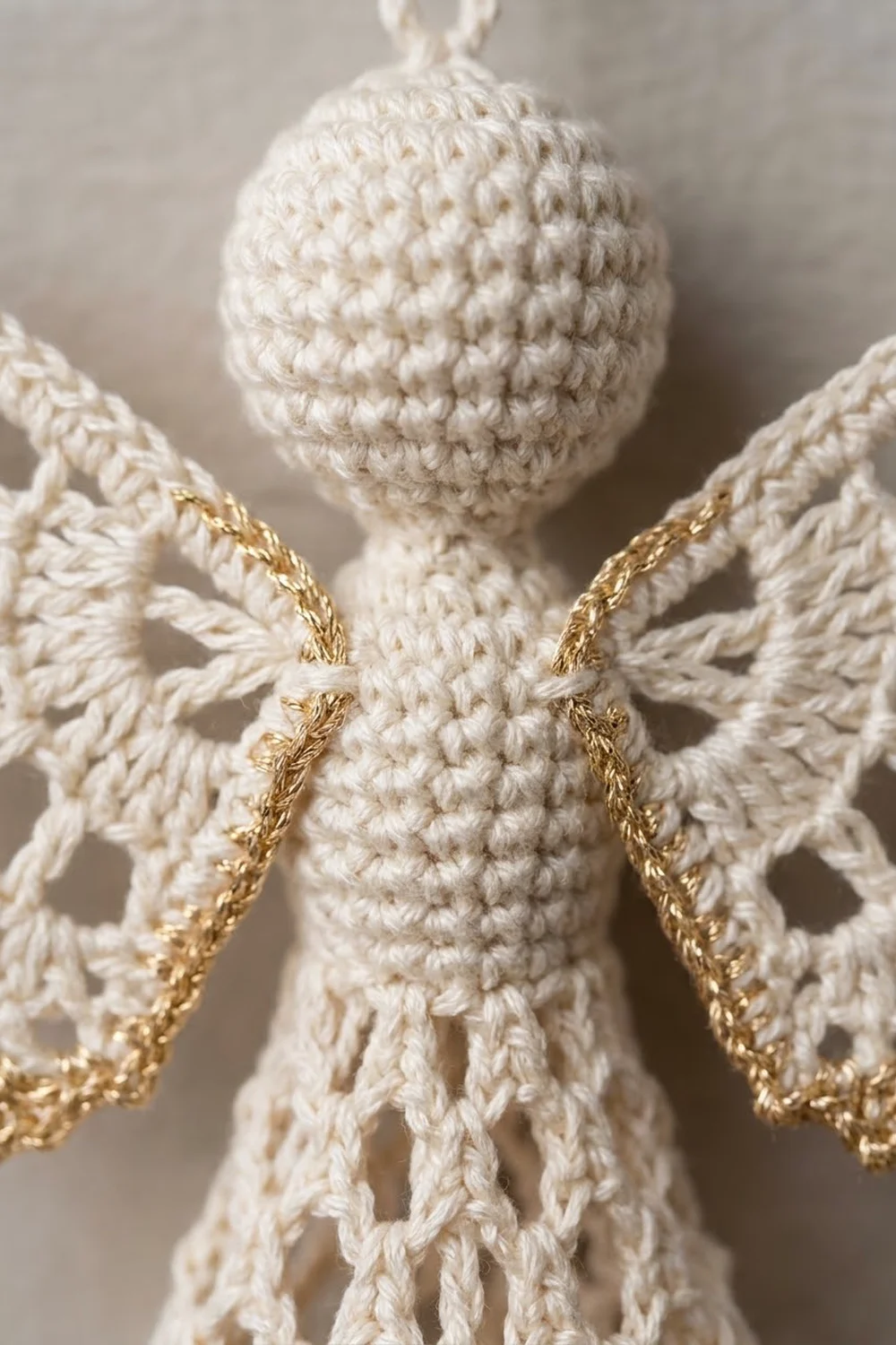 Close-up of a cream-colored crocheted angel ornament with gold-trimmed wings against a soft neutral background.