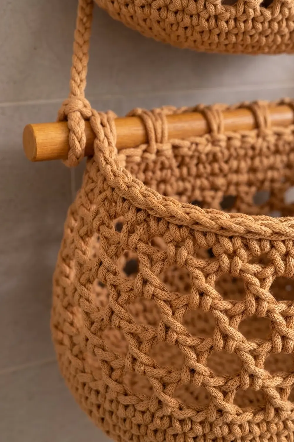 Tan crocheted wall hanging with open mesh texture and rope detail wrapped around a natural wood dowel rod.