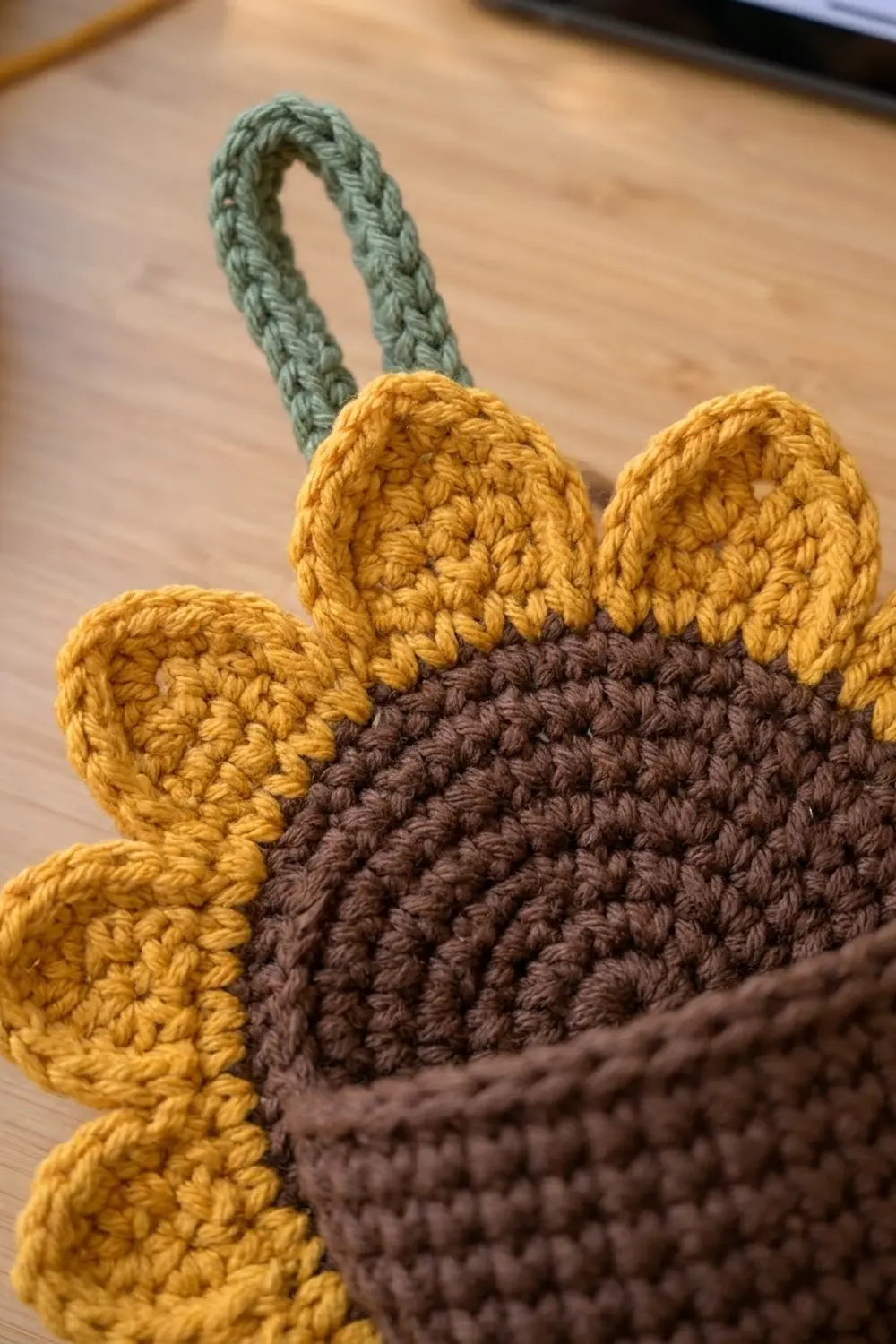 Crocheted bear with golden ears and brown body attached to a sage green braided rope handle on wood.