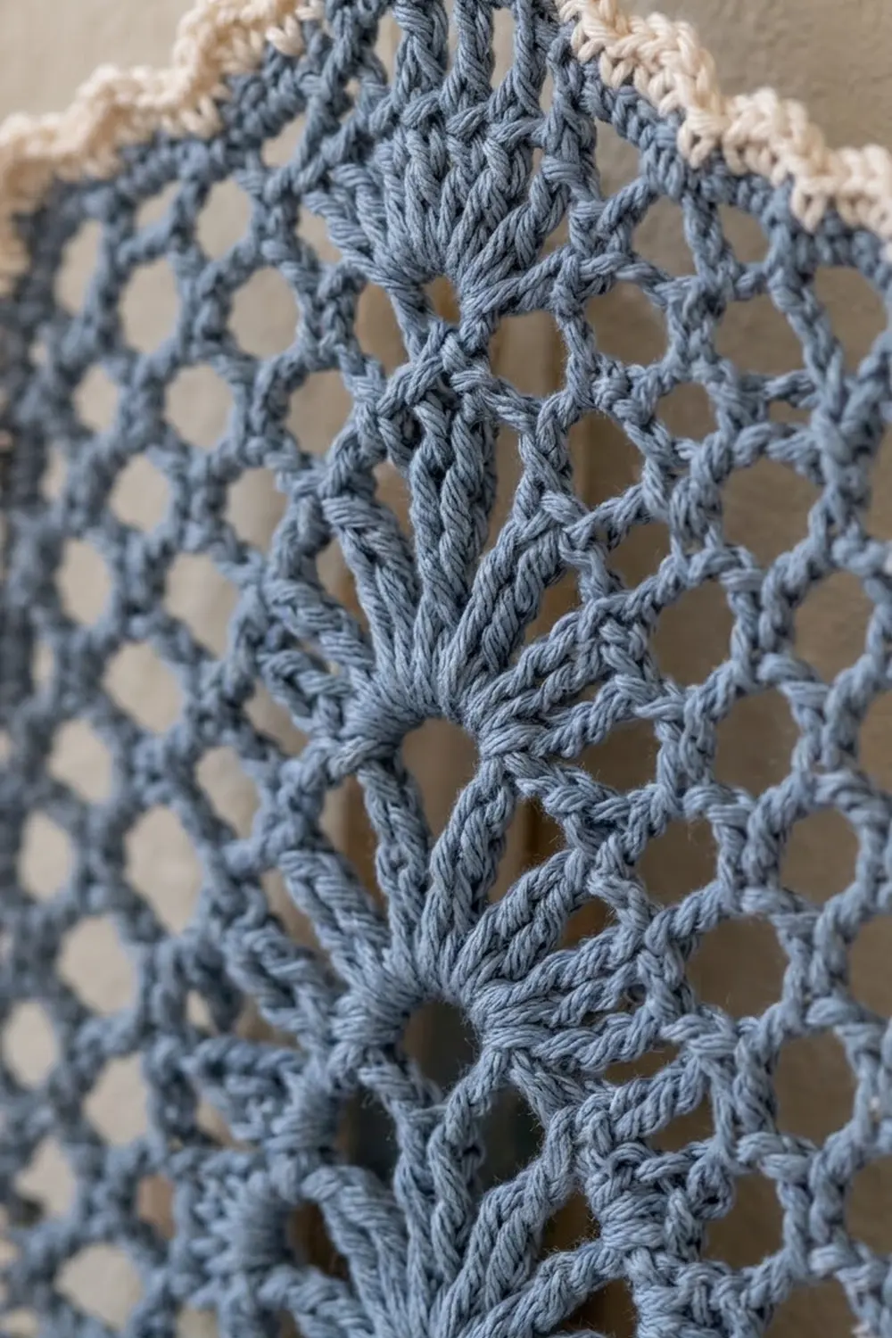 Close-up of blue-gray and white crocheted lace fabric displaying an openwork diamond pattern against a neutral background.