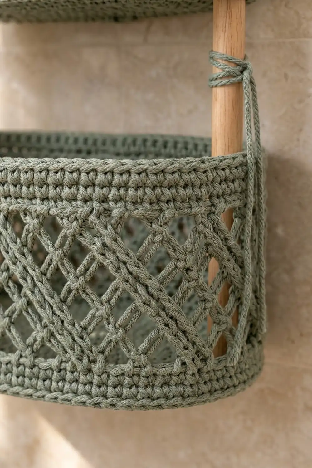 Sage green crocheted basket with open diamond lattice pattern attached to natural wooden dowel with rope knot.