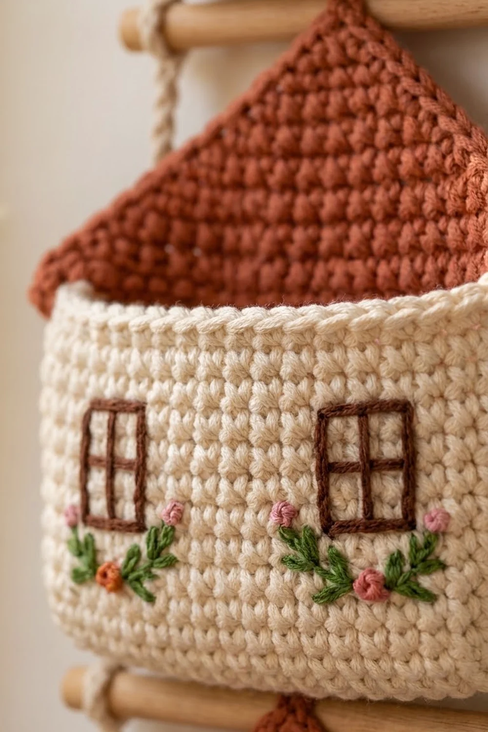 A detailed crocheted cottage featuring cream walls, embroidered windows and flowers, topped with a rust-brown roof.
