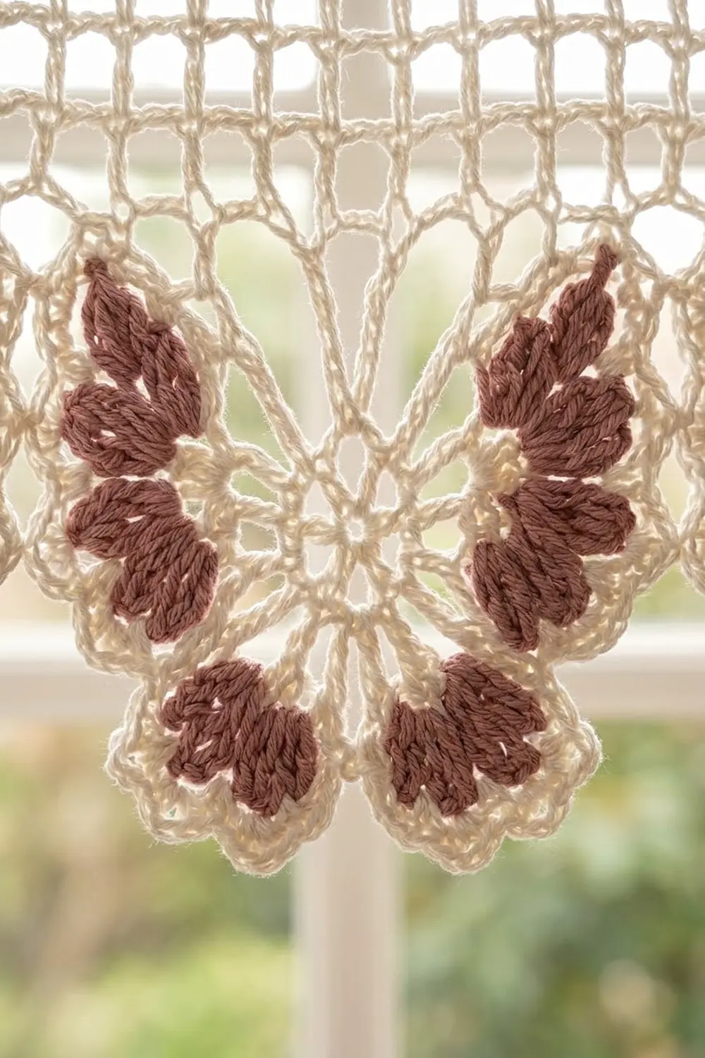 Crocheted lace curtain with beige mesh and burgundy leaf motifs hanging in a window.