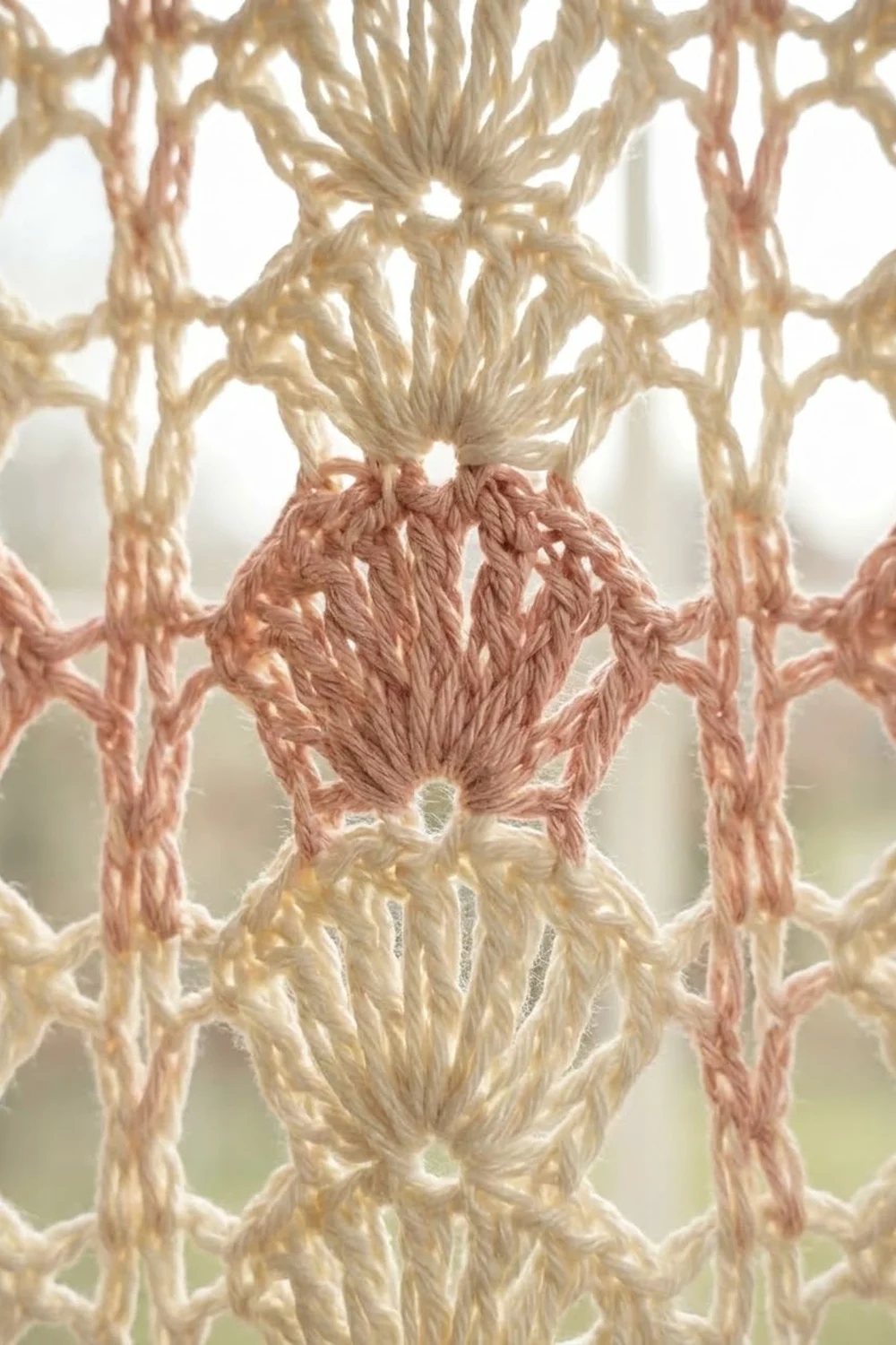 Close-up of macramé wall hanging with cream and dusty rose knotted rope forming geometric patterns against white background.