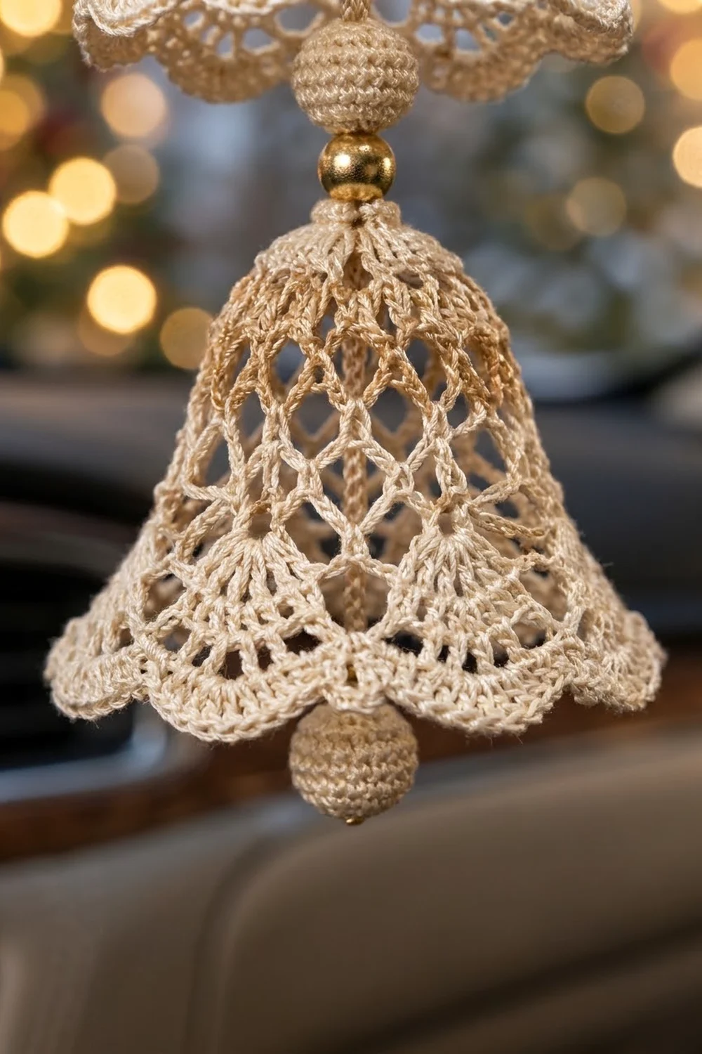 Cream-colored crocheted lace bell ornament with gold bead accent hanging against blurred holiday bokeh lights.