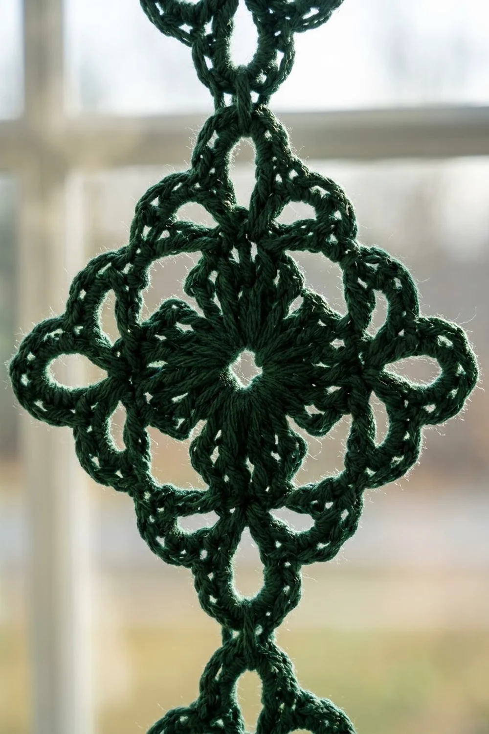 A dark green crocheted snowflake ornament with delicate lace patterns hanging in front of a blurred window.