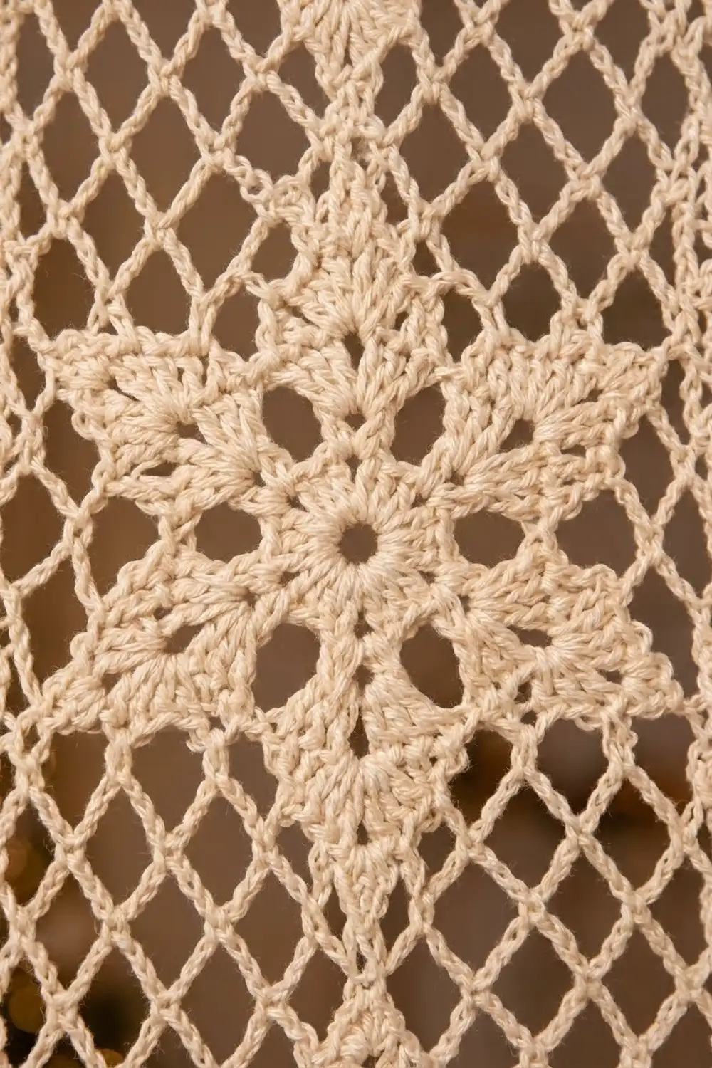 Cream-colored crocheted lace fabric with diamond-shaped openings and detailed stitching against a soft brown background.