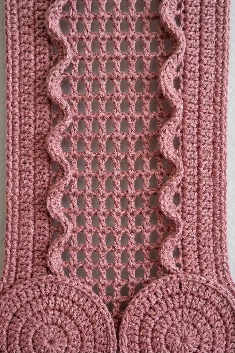 A close-up of dusty pink crocheted fabric featuring an open lace diamond pattern with textured cable borders on either side.