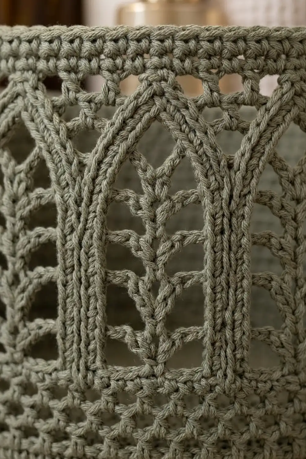 Close-up of pale sage-green crocheted fabric with diamond openwork pattern and softly blurred warm background.