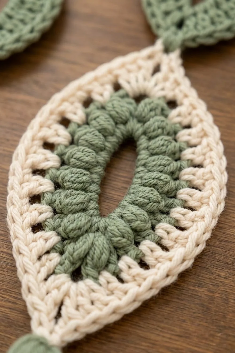 Close-up of a crocheted cowl scarf with cream border and sage green center detail on wooden surface.