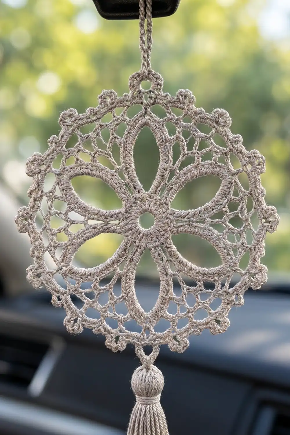 Cream-colored crocheted ornament with intricate lace pattern hanging by beige cord from car vent, blurred green foliage in background.