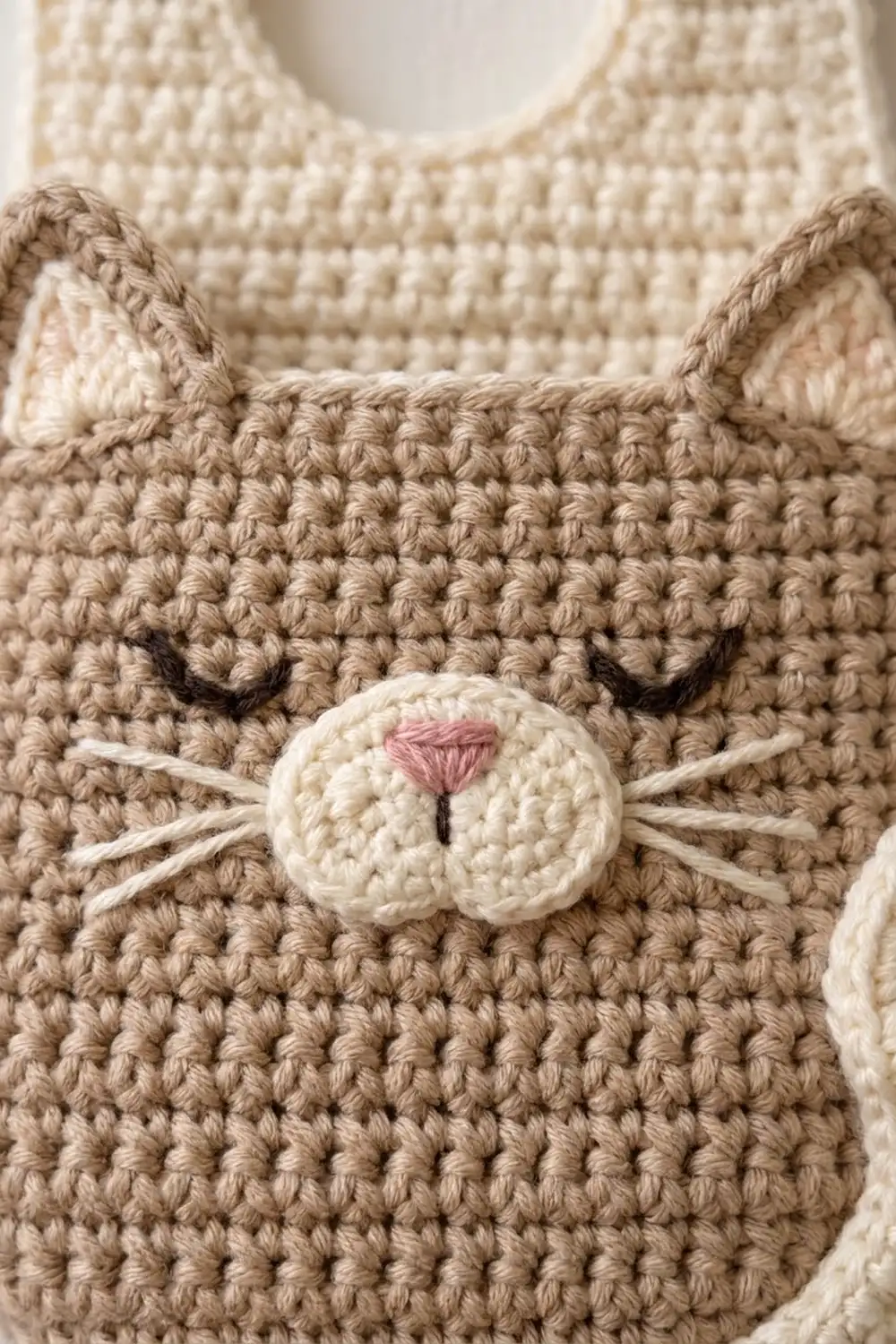 Crocheted cat face with tan ears, black eyes, pink nose, and whiskers on cream-colored knit fabric.
