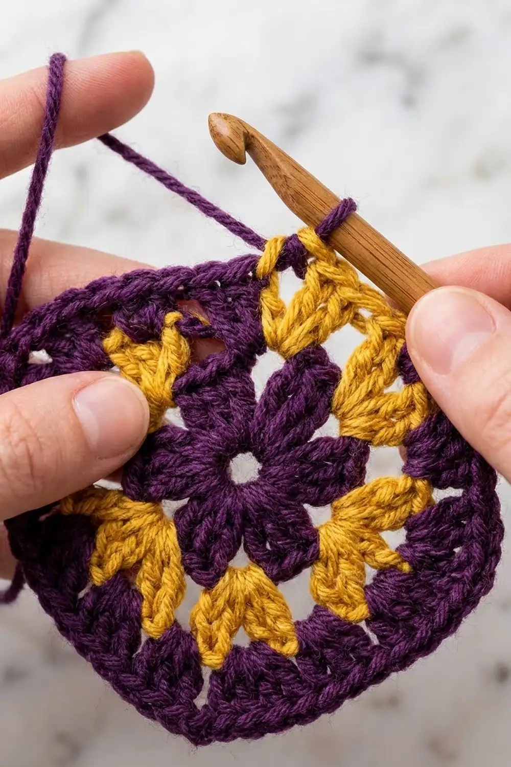Hands holding a wooden crochet hook with purple and yellow granny square work in progress.