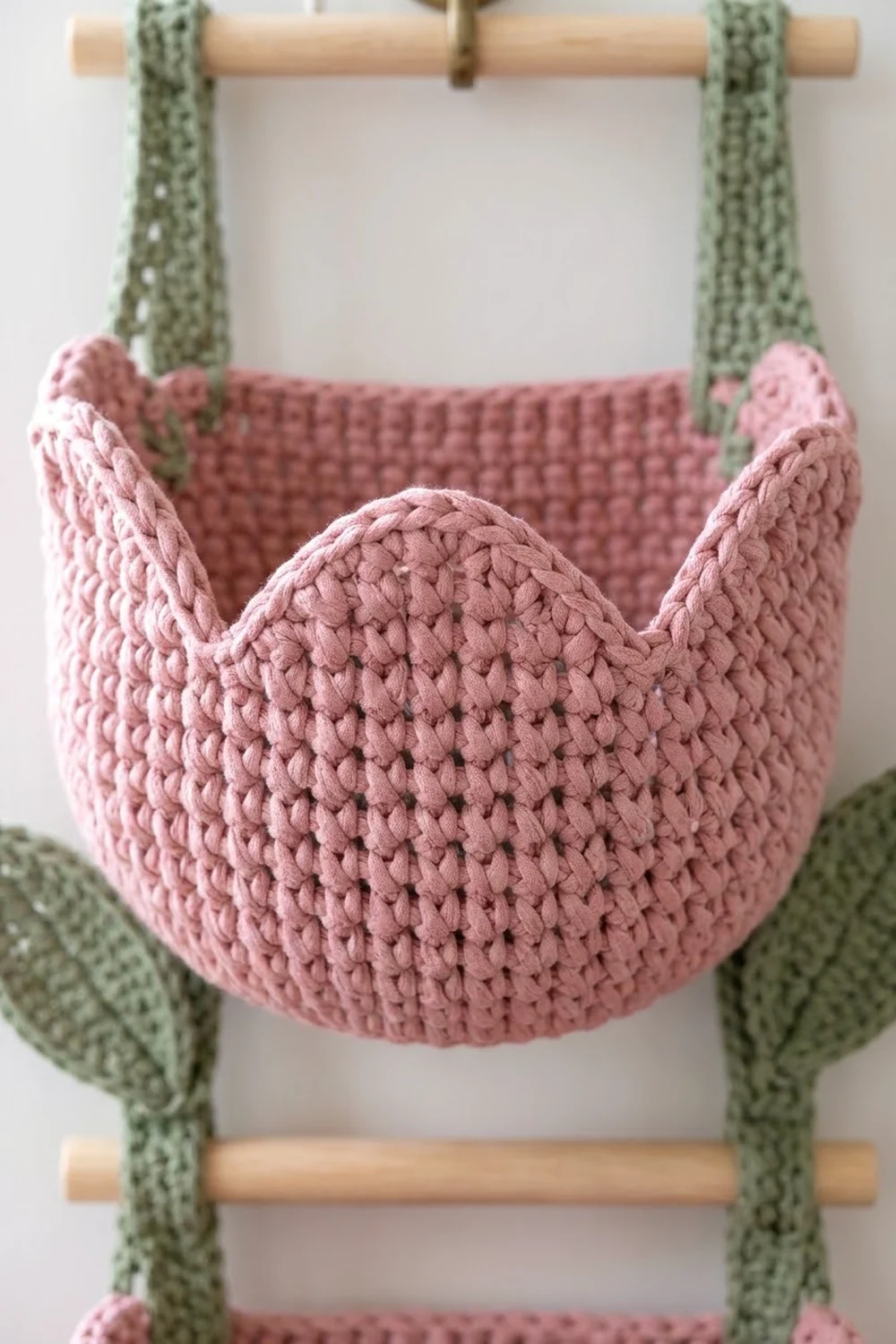 Dusty pink crocheted basket with sage green handles displayed on a natural wood rod.