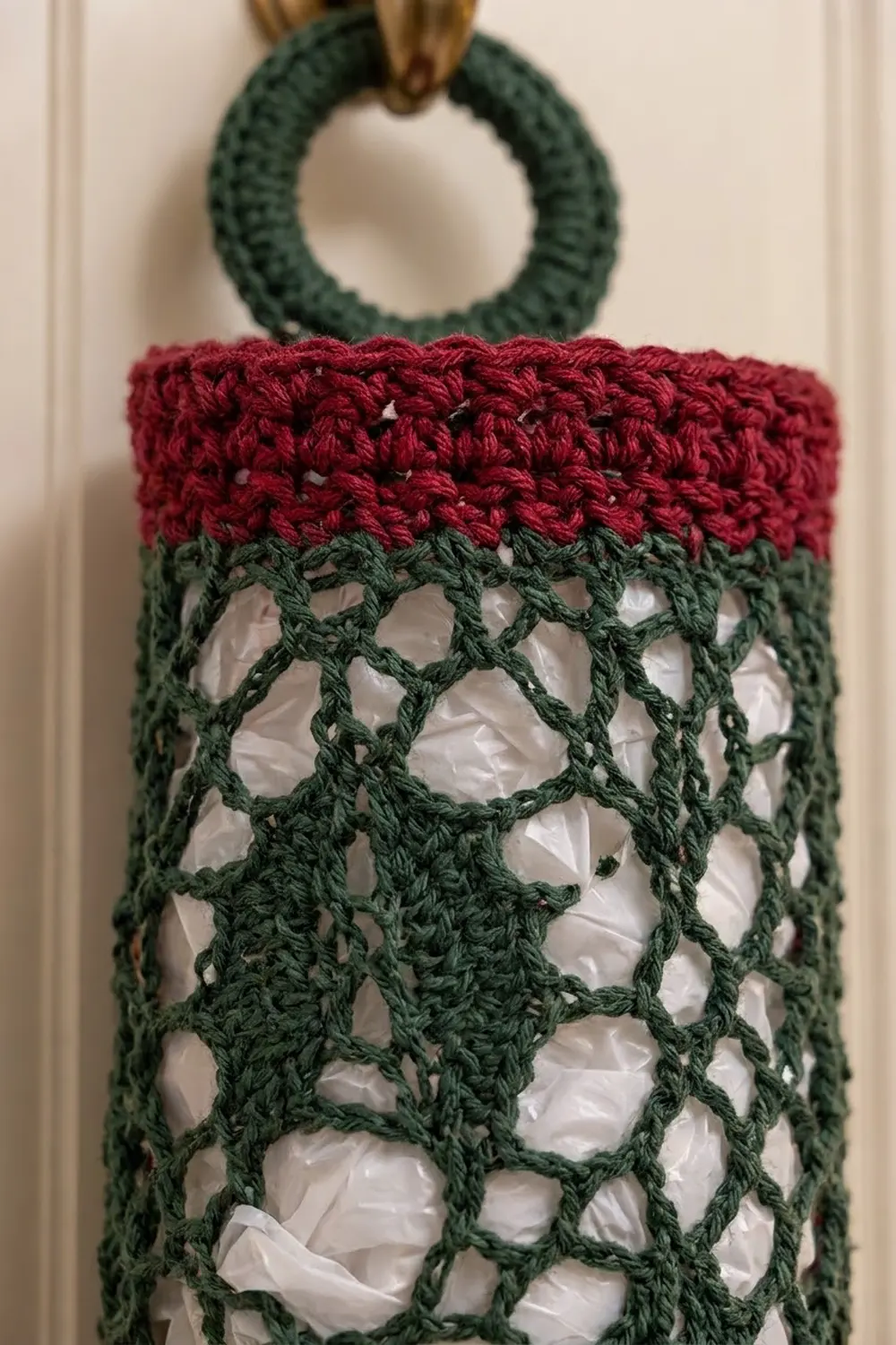 Crocheted mesh tote with dark green lattice, burgundy stripe, and matching handle held against cream wall.