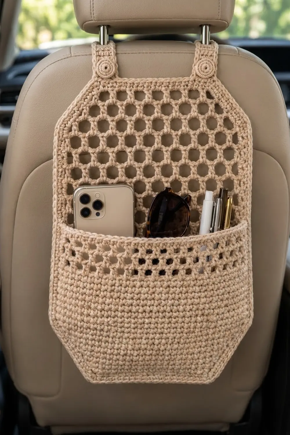 Cream crocheted car seat organizer with open-work pattern hanging on beige seat, holding phone and sunglasses.