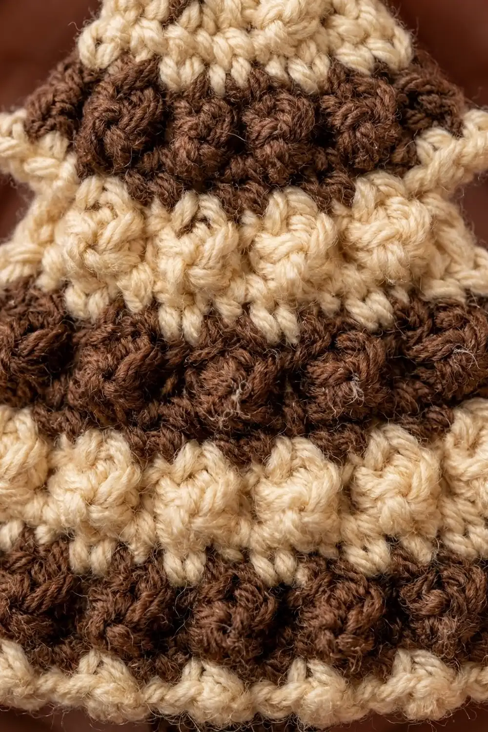 Close-up of striped crochet fabric alternating cream and brown rows, showing detailed bumpy texture against a soft brown background.