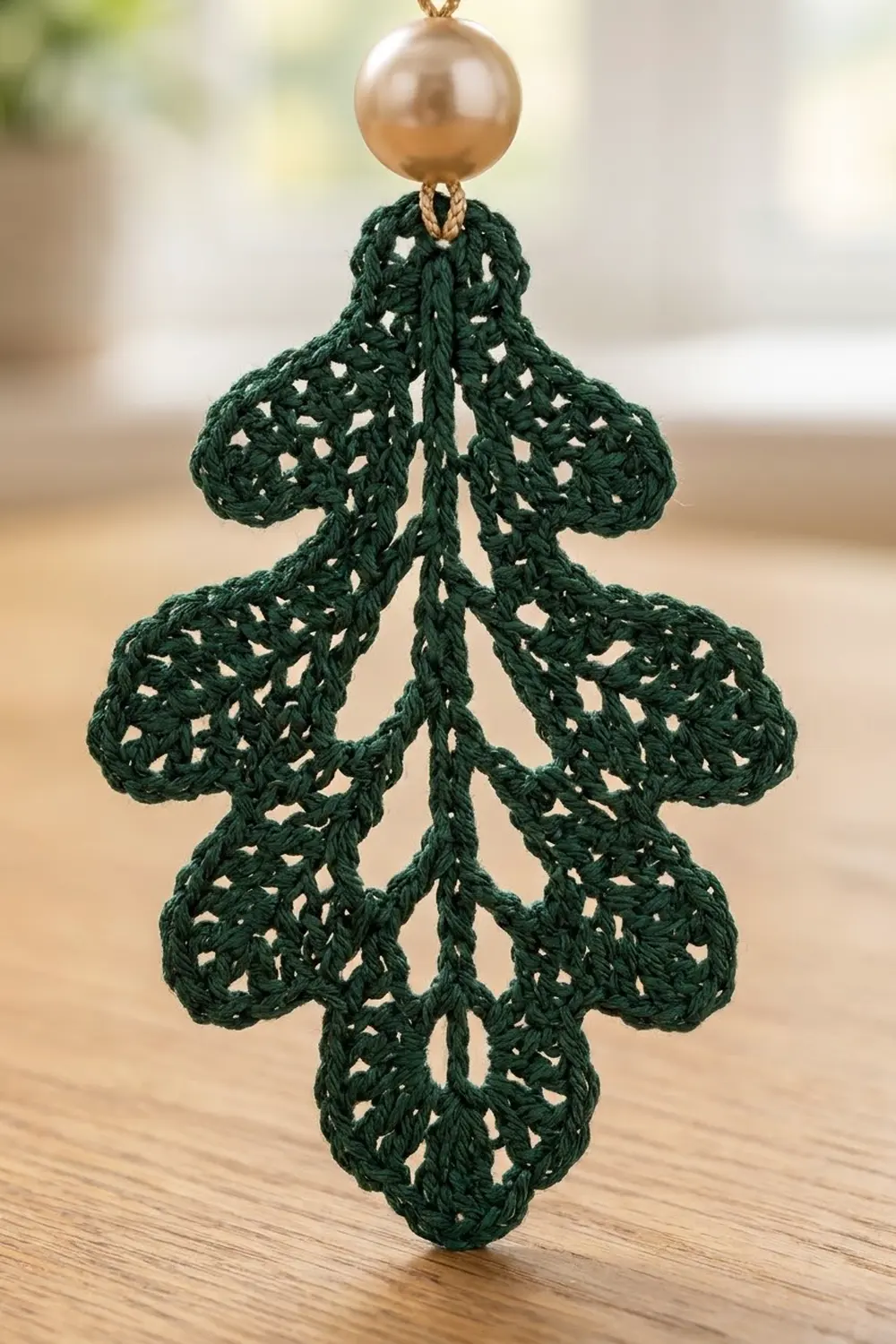 Dark green crocheted tree ornament with openwork pattern and gold ball topper, hanging against blurred background above wooden surface.