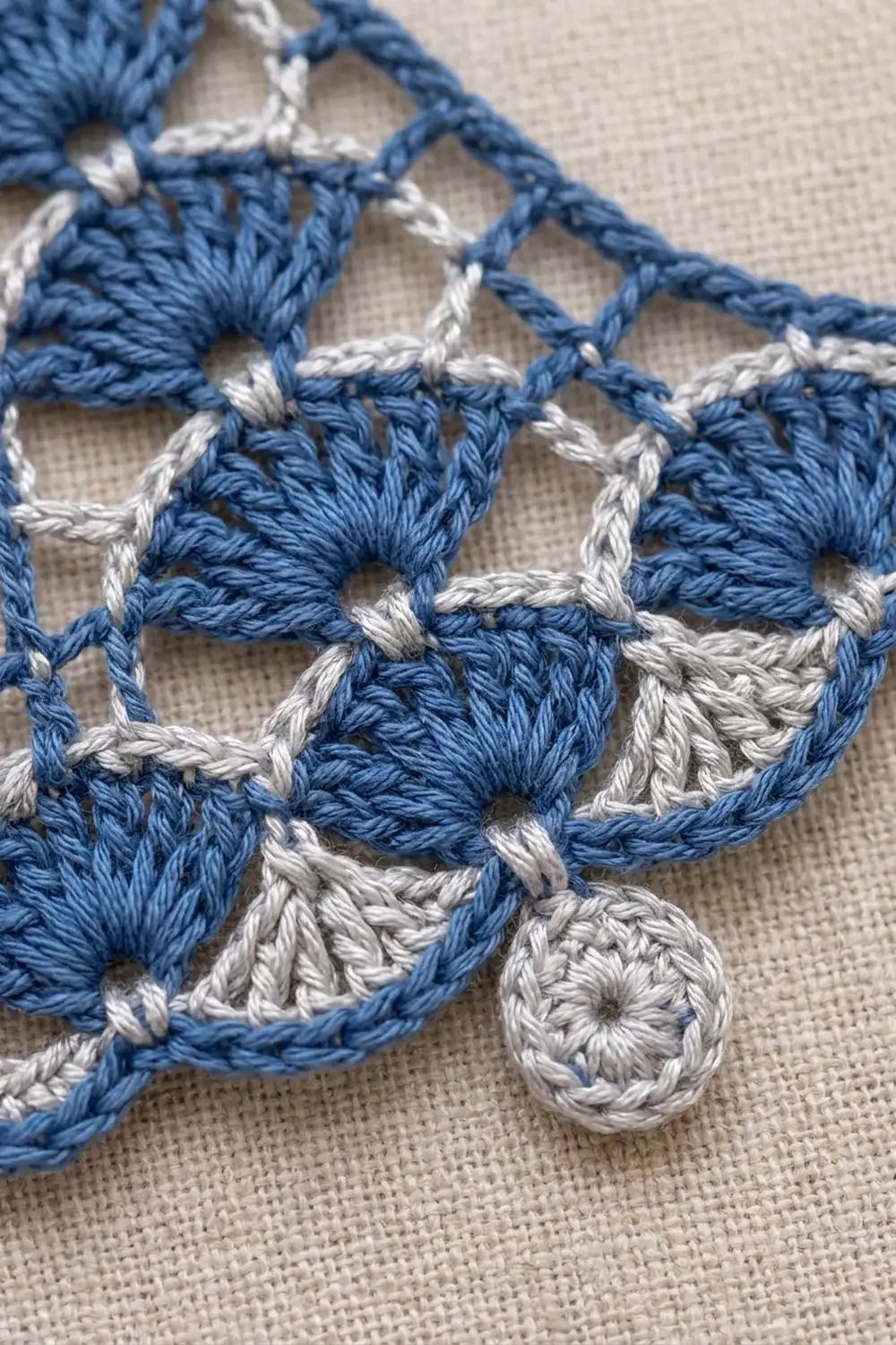 Close-up of blue and white crocheted lace trim with shell motifs displayed on beige linen fabric.
